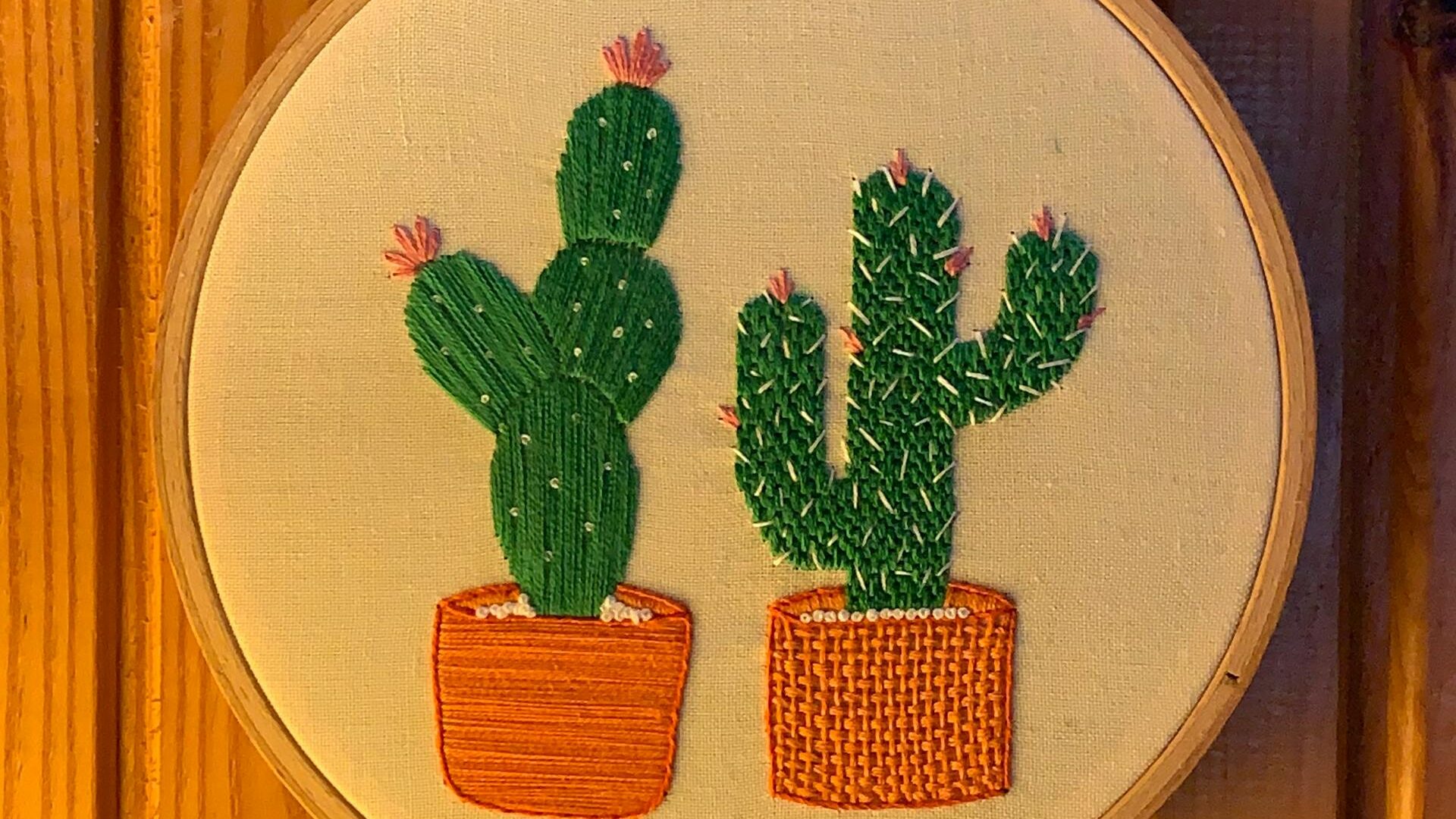 Embroidered cacti in pots, displayed in hoop on wooden wall.