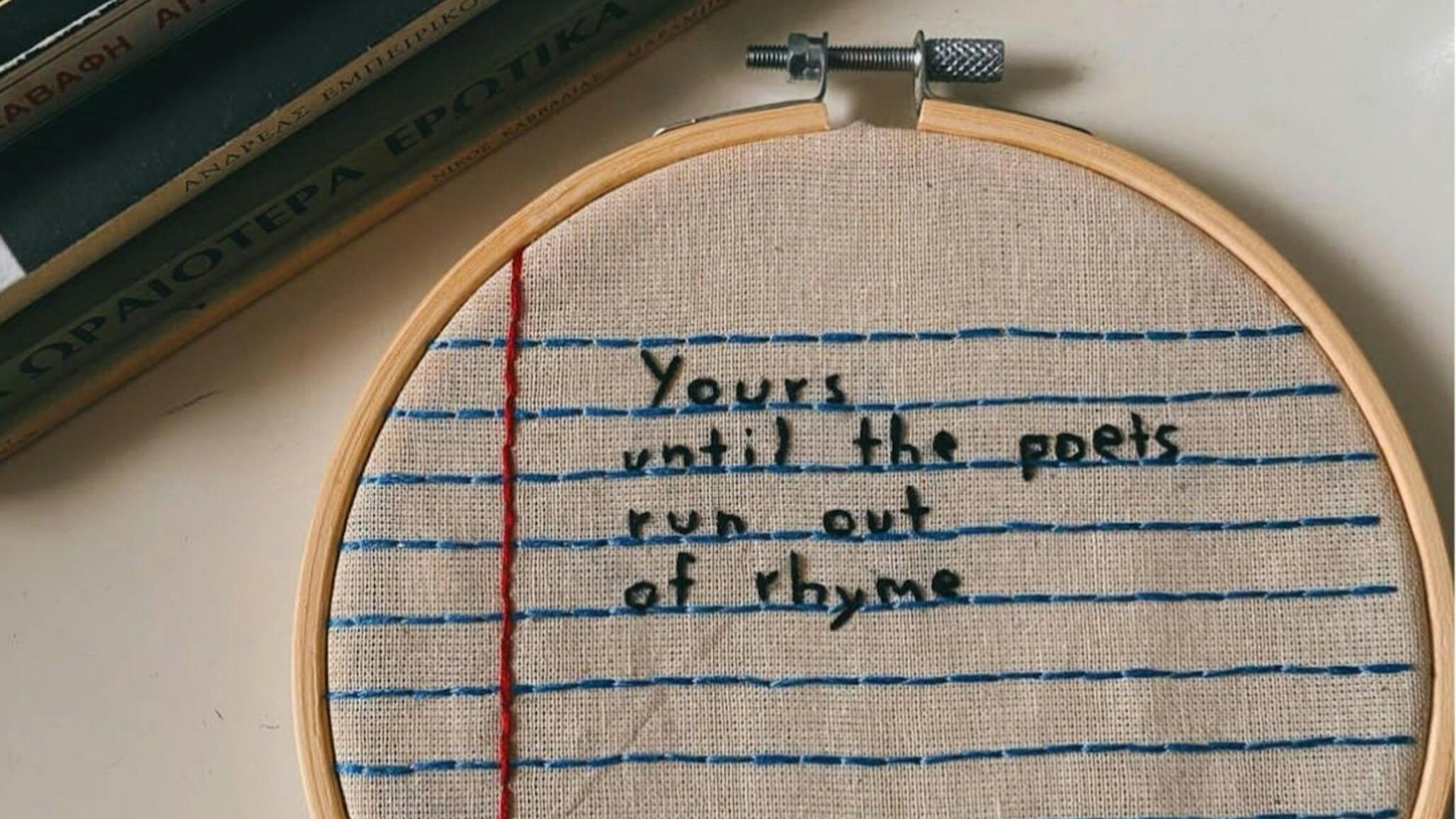 Embroidery hoop with text on notebook-style fabric, beside stacked books.