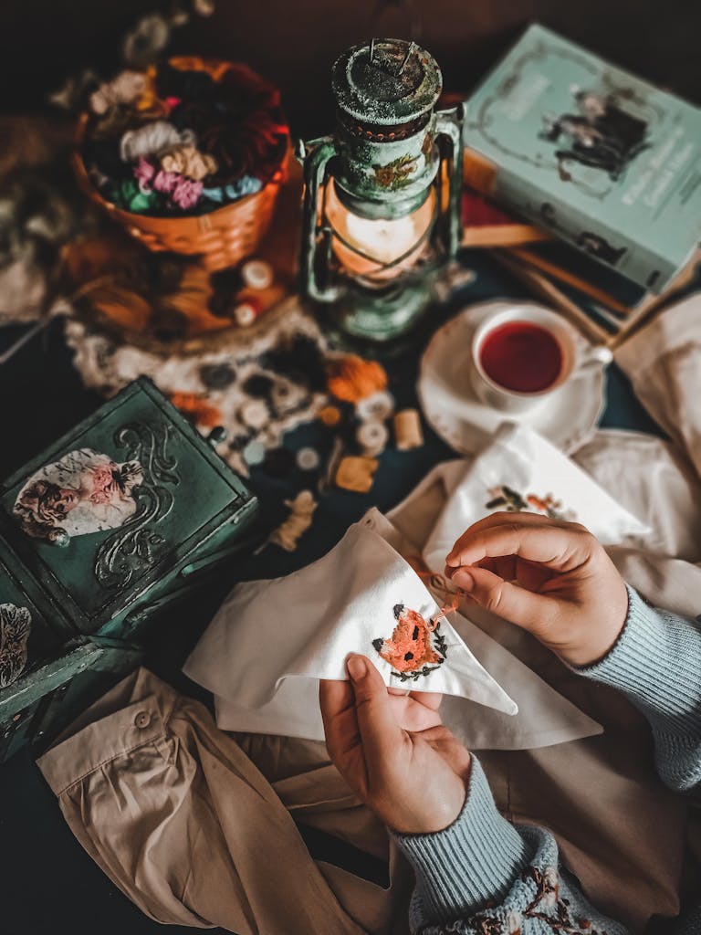 Person embroidering beside a vintage lamp with tea and books. Cozy crafting scene.